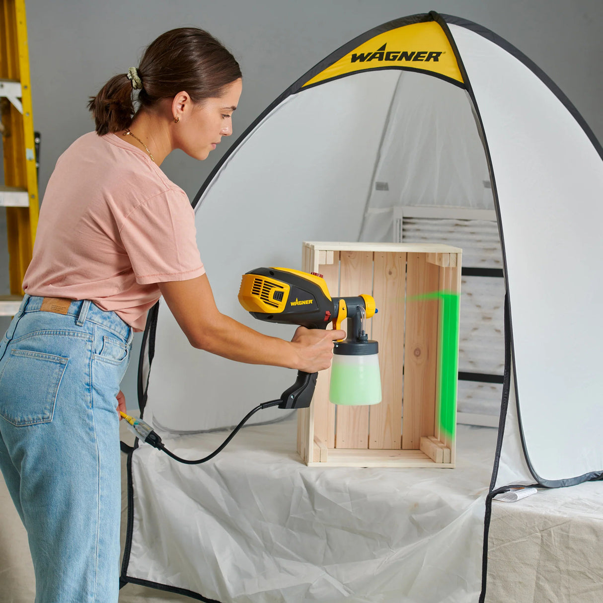 Person using a Wagner paint sprayer on a wooden crate with a spray tent in the background.