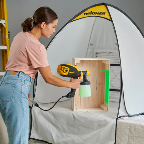 Person using a Wagner paint sprayer on a wooden crate with a spray tent in the background.