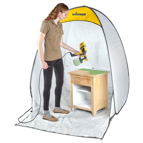 Person using a Wagner paint sprayer inside a portable spray booth to paint a wooden table.