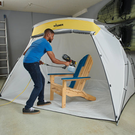 Person using a Wagner paint sprayer inside a portable spray booth to paint a wooden chair.